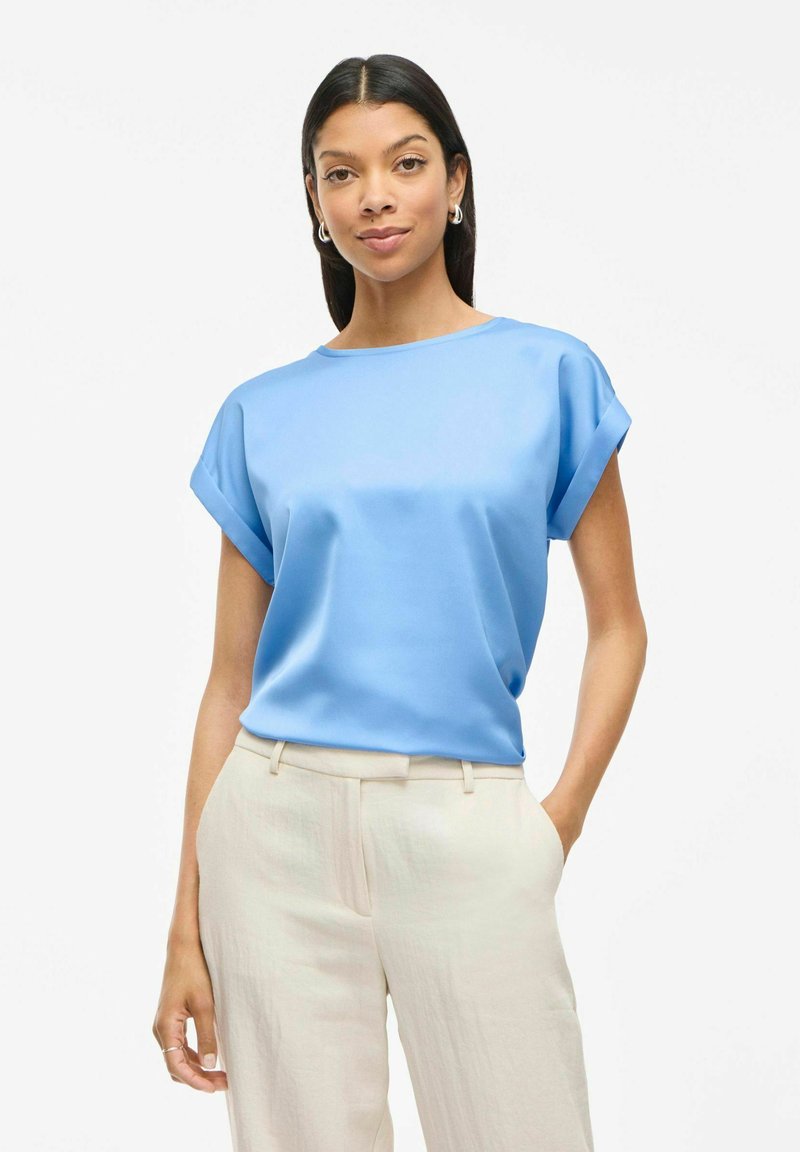 Blusa de satén azul con mangas enrolladas, cuello redondo, textura suave, combinada con pantalones de pierna ancha de color beige claro. Diseño sencillo con un atractivo clásico.