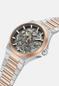 Srebrny zegarek ze stali nierdzewnej z różowym złotem na lunetce, otwartym cyferblacie ujawniającym skomplikowane mechanizmy, z ogniwowym paskiem i kanciastymi znacznikami.
