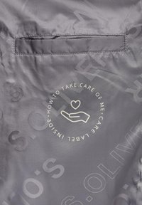 Grijs stoffen label met verzorgingsinstructies, cirkelontwerp met een hand- en harticoon, en de tekst "HOE IK TE VERZORGEN." Geweven oppervlakte.