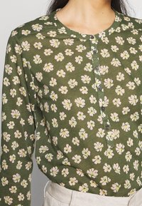 Vihreä kukkakuosiset bleiseri, joka on valmistettu kevyestä kankaasta. Siinä on pyöreä pääntie, nappi edessä ja pieniä vaaleanpunaisia kukkia keltaisilla korostuksilla.