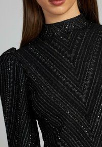 Schwarzes Pulloveroberteil mit hohem Hals, aus strukturiertem Stoff und mit horizontalen sowie Zickzackmustern und glitzernden Akzenten.
