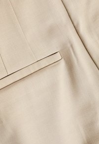 Beige bleiserikangas, jossa on sileä pinta, sivutasku ja hienovarainen kudontakuvio. Muotoiltu malli puhtailla linjoilla.