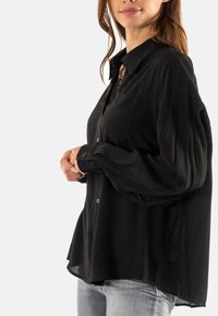 Camisa negra de botones con un bolsillo, de tela suave, corte relajado y mangas largas. Presenta un ligero brillo y un dobladillo más largo por detrás.