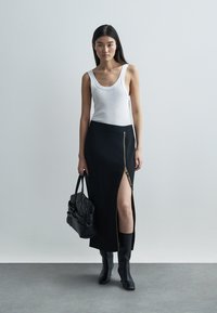 Saia preta com fecho lateral e fenda, combinada com um top branco. A modelo segura uma mala preta e usa botas pretas até ao joelho.