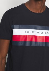 Námořnické bavlněné tričko s vodorovnými pruhy v tmavě modré, bílé a červené barvě. Uprostřed je logo s nápisem "TOMMY HILFIGER" v bílé barvě. Hladká textilie.