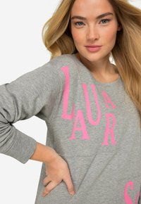 Grå sweatshirt med stora rosa bokstäver som stavar "LAURAS". Den har rundad halsringning och långa ärmar med avslappnad passform.