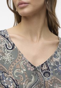 V-neck vaatteessa on paisley-kuvio, jossa on hillittyjä vihreitä, vaaleanpunaisia ja sinisiä sävyjä. Se on valmistettu pehmeästä ja kevyestä kankaasta, jossa on hienovarainen kiilto.