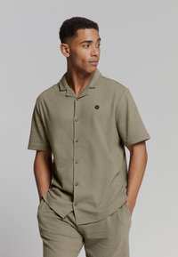 Khaki kortekogels met een kraag en zes knopen, met een klein zwart hexagonaal logo aan de linkerzijde. Zacht, gestructureerd stof.