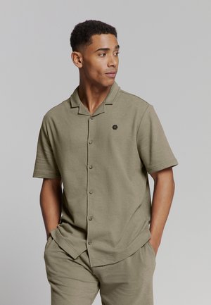 Khaki kortekogels met een kraag en zes knopen, met een klein zwart hexagonaal logo aan de linkerzijde. Zacht, gestructureerd stof.
