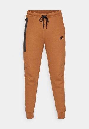Nike Jogginghose in einem warmen Braunton, mit einem schwarzen Logo, elastischem Bund, seitlicher Reissverschlusstasche und schmal zulaufenden Beinabschlüssen.
