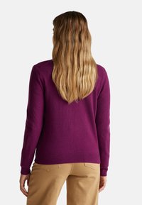 Fialový svetr vyrobený z měkkého materiálu, s obdélníkovým výstřihem, dlouhými rukávy a žebrovanými lemy; spárován se světle hnědými kalhotami.