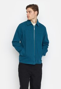 Teal zip-up jack met twee voorzakken, een kraag, en met knopen afgewerkte manchetten, gemaakt van een textuurstof. Draagt over een licht shirt.