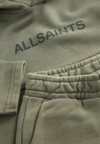Completo con felpa verde oliva e pantaloni tuta dal texture morbida. La felpa presenta il testo "ALLSAINTS", mentre i pantaloni hanno una vita elastica e cuciture sottili.