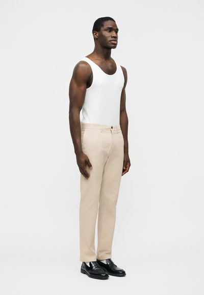 Άνδρας που στέκεται φορώντας λευκό αμάνικο πουκάμισο, μπεζ παντελόνι και μαύρα επίσημα παπούτσια, στραμμένος ελαφρώς προς τα δεξιά σε απλό φόντο.