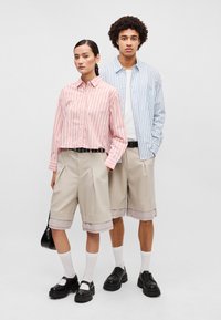 Kaks mudelit kannavad nööpidega särke, millel on vertikaalsed triibud: üks roosa ja punane, teine sinine ja valge. Mõlemal on seljas beežid šortsid ja mustad kingad.