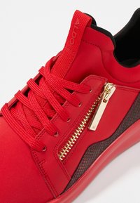 Červená sportovní teniska s texturovanými stranami, plochými tkaničkami a zlatým zipovým detailem. Disponuje hladkým syntetickým materiálem a černým vnitřním podšitím.