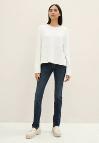 Bílý pletený svetr s žebrovanými manžetami, v kombinaci s tmavě modrými džínami. Model nosí béžové platformové boty. Jednobarevné pozadí.