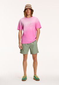 Világos rózsaszín gradiens T-shirt, rajta a "TRANQUILO" felirattal, olajzöld rövidnadrággal és zöld cipőkkel, sárga fűzőkkel; szalma vödörkalap.