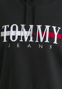 Sweat à capuche noir avec texte brodé blanc "TOMMY JEANS" sur des rayures horizontales bleu marine, blanc et rouge, avec des cordons noirs pendants.