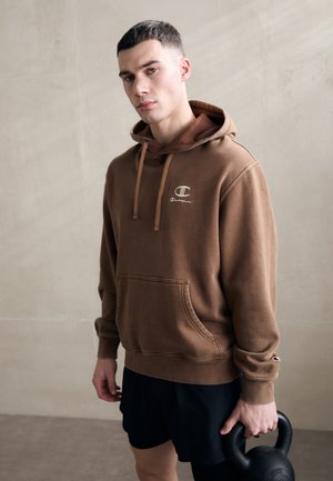 Hoodie marron en tissu doux, avec une poche frontale, des cordons de serrage et un logo sur la poitrine. Accompagné de shorts de sport noirs.