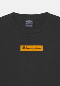 Maglietta nera con collo rotondo, caratterizzata da una toppa rettangolare arancione con la scritta "Champion" in testo corsivo nero.