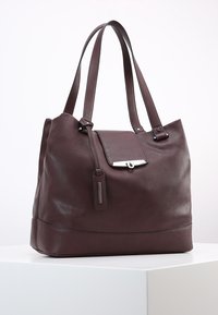 Large brown leather tote bag with smooth texture, dual shoulder straps, front flap, and metallic hardware detail.