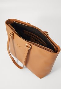 Tan leren tote bag met een ritssluiting, voorzien van gouden hardware, een interne zak en een gladde textuur. Vlakke basis voor stabiliteit.