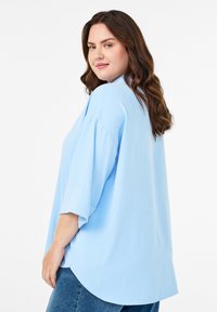 Curly brown-haired woman in light blue oversized shirt and blue jeans, smiling slightly, facing left with a white background.