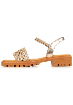 Sandalo con punta aperta con ampia fascia metallica oro intrecciata, cinturino metallico alla caviglia e suola in gomma robusta color camoscio con tacco basso a blocco.