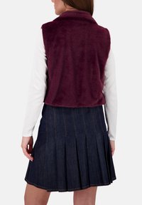 Burgunderfarbener Kunstpelz-Westen mit Kragen, getragen über einem weißen Langarmshirt. Kombiniert mit einem dunkelblauen, plissierten Rock aus Denim.
