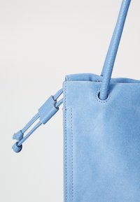 Sac fourre-tout en daim bleu clair avec un design structuré, des accents en cuir lisse, une fermeture à cordon et une poignée unique pour le transport.