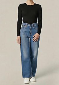 Personne portant une chemise noire à manches longues, un jean bleu taille haute à jambes larges et des chaussures blanches, debout devant un fond uni.
