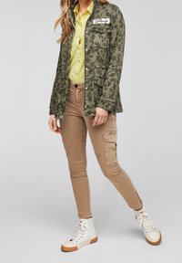 Giacca verde con motivo mimetico, dotata di bottoni a pressione e tasche, abbinata a una camicia gialla con bottoni e pantaloni marrone chiaro. Sneakers bianche alte.