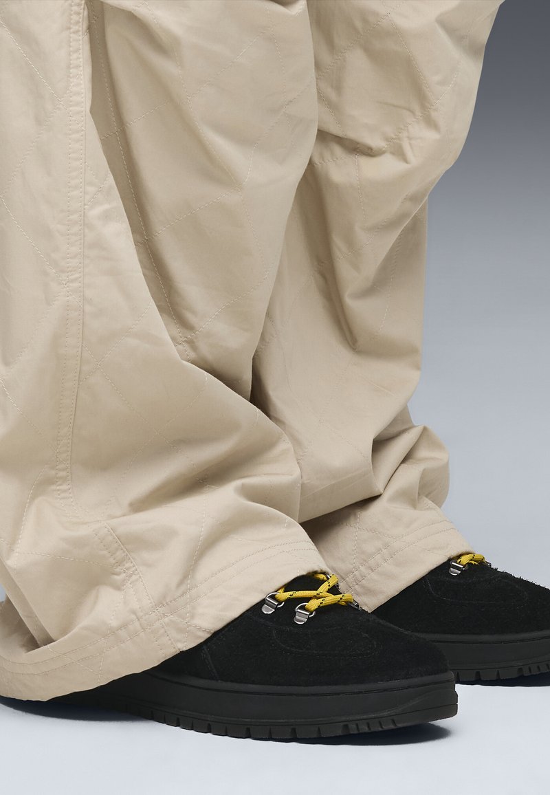 Beige, gevoerde broeken met een losse pasvorm, gecombineerd met zwarte schoeisel met gele veters en zilverkleurige accenten, tegen een grijze achtergrond.