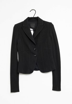 Czarna dopasowana marynarka z klapami i dwoma przednimi guzikami zawieszona na czarnym wieszaku na białym tle.