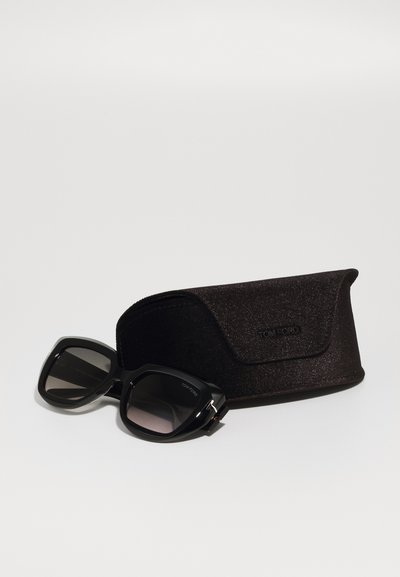 Occhiali da sole neri Tom Ford con lenti sfumate, montatura quadrata e aste spesse, accanto a un astuccio nero testurizzato con chiusura magnetica.