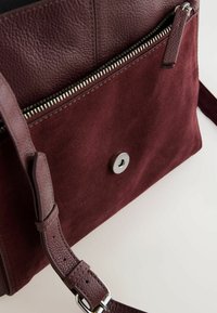 Borsa bordeaux con parte superiore in pelle liscia, corpo in suede, zip argentata e chiusura a scatto. Presenta una tracolla regolabile e una finitura texturizzata.