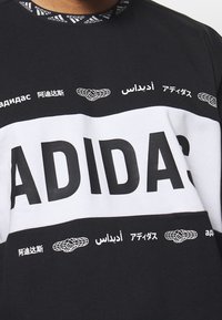 Czarna i biała bluzą Adidas z wyraźnym napisem "ADIDAS", wielojęzycznym brandingiem i wzorzystym kołnierzykiem. Wykonana z materiału bawełnianego, w standardowym kroju.