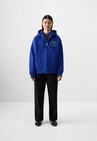 Blå zip-up hoodie med framfickor, broderad design på bröstet, i kombination med svarta lösa byxor och mörka skor.