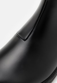 Botín de cuero negro hasta el tobillo con un acabado suave, diseño minimalista y un pequeño fuelle en la parte delantera. Detalles de costura a lo largo de las costuras.