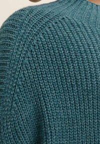 Pull en maille coloris sarcelle avec une finition texturée. Le tissu présente un motif de mailles serrées et lâches le long de l'encolure et des épaules.