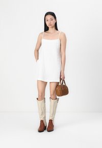 White linen dress with shoulder straps, brown leather and beige cowboy boots, and a small brown handbag with a textured pattern.