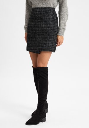 Schwarzer Tweed-Minirock mit diagonaler Wickel-Optik, ausgestattet mit einer strukturierten Oberfläche und kombiniert mit schwarzen Overknee-Stiefeln aus Wildleder.
