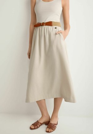 Kobieta w beżowym topie bez rękawów, beżowej spódnicy midi z wysokim stanem i brązowym paskiem oraz brązowych sandałach na obcasie, stoi przy białej ścianie.