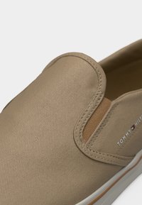 Chaussure en toile beige à enfiler avec un panneau latéral élastique et une semelle blanche, avec le logo Tommy Hilfiger sur le côté.