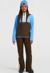Blauwe en bruine fleece pullover met hoge kraag en halve rits, gecombineerd met bruine broek met crèmekleurige accenten aan de zijkanten. Zwarte sneeuwlaarzen.