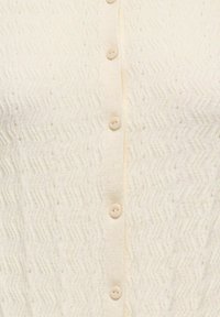 Detailní záběr krémově zbarvené pleteniny se vzorem cikcak a pěti shodnými kulatými knoflíky seřazenými vertikálně.