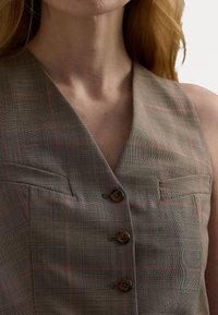 Brun rutig väst med V-ringning, som har fyra bruna knappar och två bröstfickor med subtila orangea accenter i mönstret.