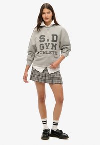 Grå sweatshirt med sort trykt tekst, lagd oven på en hvid skjorte med krave, parret med en ternet plisseret nederdel og sorte sko med stribede sokker.