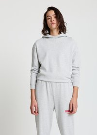 Sweat à capuche gris clair avec une texture douce et une coupe ample, doté d'une poche avant et d'une capuche à cordon, associé à un pantalon de survêtement gris assorti avec un texte rouge.